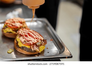 chef adding special sauce from a plastic squeeze bottle to a gourmet bacon cheeseburger with melted cheese, bacon strips, tomatoes, and pickles on a toasted bun