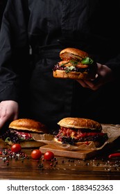 Chef In A Black Apron Holding A Juicy Burger, With A Table Standing In Front Of Him, Hands Only Visible