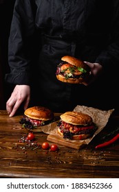 Chef In A Black Apron Holding A Juicy Burger, With A Table Standing In Front Of Him, Hands Only Visible