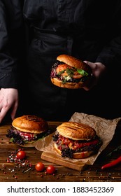 Chef In A Black Apron Holding A Juicy Burger, With A Table Standing In Front Of Him, Hands Only Visible