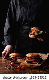 Chef In A Black Apron Holding A Juicy Burger, With A Table Standing In Front Of Him, Hands Only Visible