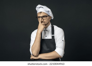 A Chef In A Black Apron On A Black Background Adjusts His Glasses. Smart Guy Concept.