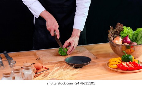 chef in black apron slices a green bell pepper on wooden cutting board, surrounded by fresh vegetables and ingredients - Powered by Shutterstock