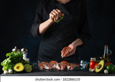 The chef in black apron sprinkle lime juice on salmon steak. Professional kitchen table on black background for design. Traditional recipe of preparing fish. Adding spices for taste. Food concept.