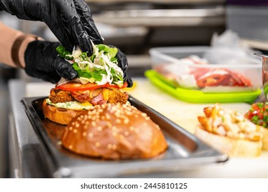 A chef in black gloves assembles a gourmet burger with fresh toppings on a bun, in a professional kitchen setting. The burger consists of lettuce, tomatoes, and other fresh ingredients. - Powered by Shutterstock