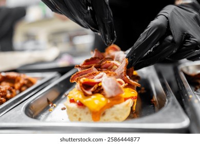 A chef in black gloves skillfully layers sliced meats over melted cheese on a biscuit in a restaurant kitchen. The image captures the artistry of food preparation and presentation.