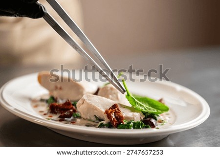 A chef in black gloves uses precision tweezers to plate a gourmet chicken dish featuring seared medallions, fresh spinach, and sun-dried tomatoes in a creamy sauce on a white oval plate