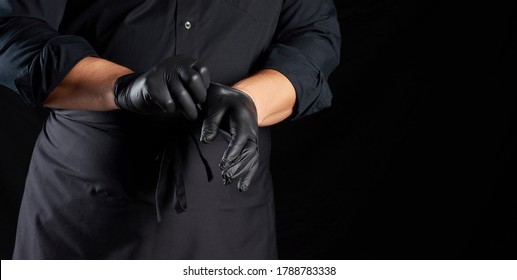 chef in black shirt and apron puts black latex gloves on his hands before preparing food, black background, copy space