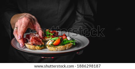 chef in a black suit holds in his hands plate with Bruschetta on Dark grey black background