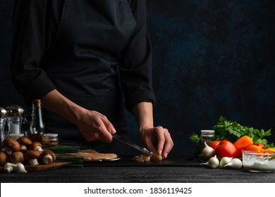 The chef in black uniform cuts with knife the chicken fillet on the professional kitchen on dark background. Backstage of cooking dinner with meat, mushrooms, vegetables and spices. Food concept. - Powered by Shutterstock