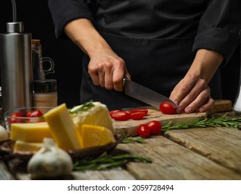 A chef in a black uniform is cutting tomatoes on a cutting board. Cheese, garlic, spices, rosemary sprigs. Cooking salad, sauce, pizza, focaccia. Close-up. Wooden texture.