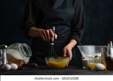 The Chef In Black Uniform Mixed All Ingredients For Pie Isolated On Dark Background. The Chef's Hands With Corolla Preparing The Meal. Recipe From The Book. Food Concept.