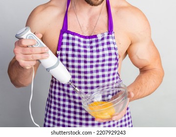 Chef bodybuilder in protective apron uses a blender to beat scrambled eggs on whie kitchen table. Muscular man cooking.