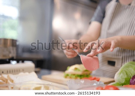 Chef carefully places a slice of ham onto a sandwich in preparation, surrounded by fresh vegetables on the counter.