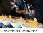 A chef carefully uses a culinary torch to caramelize the tops of freshly prepared lemon tart slices in a modern kitchen.