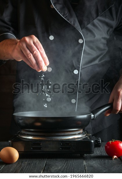 Chef or cook adds\
salt while cooking eggs in a pan. Work environment on vintage\
kitchen table. Vertical\
image