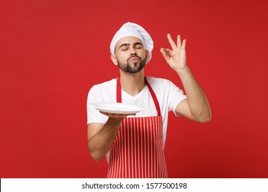 Chef Cook Or Baker Man In Striped Apron Toque Chefs Hat Isolated On Red Background. Cooking Food Concept. Mock Up Copy Space. Hold Empty Blank Plate With Place For Food Making Okay Taste Delight Sign