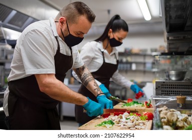 Chef and cook with face masks cutting vegetables indoors in restaurant kitchen, coronavirus concept. - Shutterstock ID 2107622954