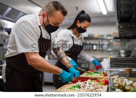 Chef and cook with face masks cutting vegetables indoors in restaurant kitchen, coronavirus concept.