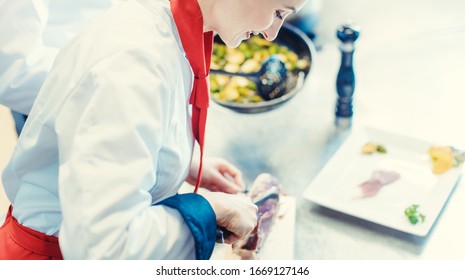 Chef Cook In Fancy Restaurant Kitchen Slicing Ham For A Dish With Plate Of Food In The Background