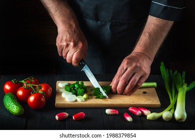 Chef or cook is hands close-up cuts young green onions on a restaurant kitchen cutting board for salad. Vegetable diet or snack idea.