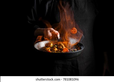 Chef Cooking Vegetables On A Pan With Fire Flambe. Black Background For Copy Text.