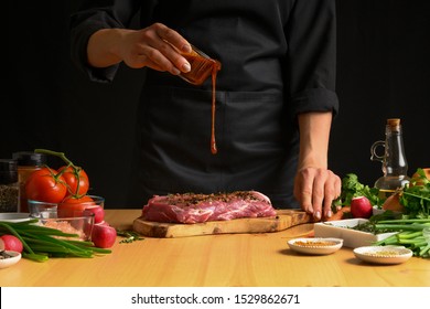 Chef Cooks Pork Steak. Pouring Sauce, Marinade On A Background With Vegetables. Recipe Book, Cooking, Restaurant Business