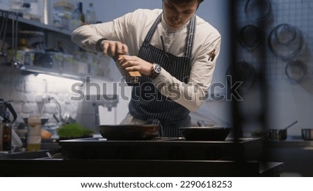 Chef cooks tasty meal. Male cook adds salt, pepper or other spices to finished or cooking dish using spice grinder. Restaurant with professional gourmet cuisine. Process of cooking. Public catering.