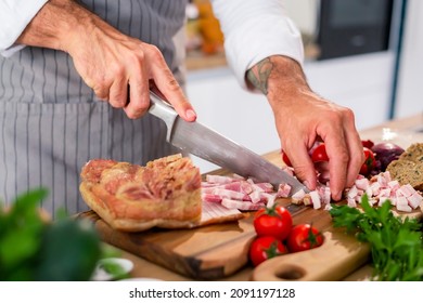 The Chef Cuts Bacon In The Kitchen On A Wooden Board. There Are Vegetables, Slices Of Bread, Spices And Small Kitchen Bowls Around The Work Surface.