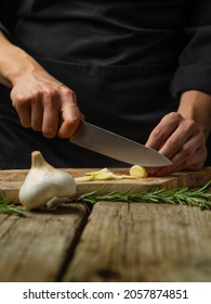 The chef cuts the garlic on a cutting board. The decor is a sprig of rosemary. Wooden texture. aromatic seasoning for different dishes - salad, soup. lasagna, sauces. Restaurant, cafe, home cooking.