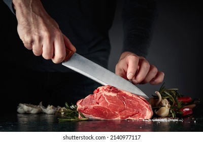 Chef cutting raw beef meat. On a table meat with rosemary, garlic, salt, and pepper.