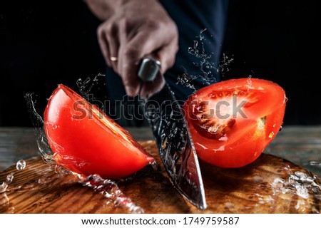 Chef cutting red tomato in half with a large knife in motion, splashes of water and juice fly in different directions and freeze in the air, shallow depth of field, selective focus, concept of cooking