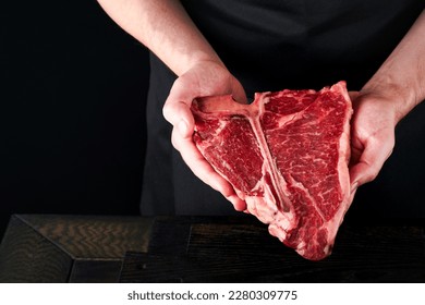 Chef cutting steak beef. Mans hands hold raw steak T-Bone on rustic wooden cutting board on black background. Cooking, recipes and eating concept. Selective focus.