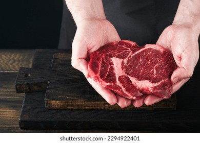 Chef cutting steak beef ribeye. Mans hands hold raw steak Ribeye on rustic wooden cutting board on black background. Cooking, recipes and eating concept. Selective focus. - Powered by Shutterstock