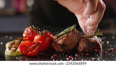 Chef decorating a plate with freshly grilled meat . Cooker adding a piece of rosemary with tweezers on top of steak - food art gourmet dinner
