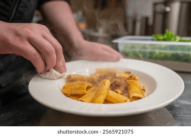 A chef elegantly garnishes a steaming plate of rigatoni pasta in a professional kitchen, showcasing culinary skills and attention to detail. Fresh herbs in the background add contrast. - Powered by Shutterstock