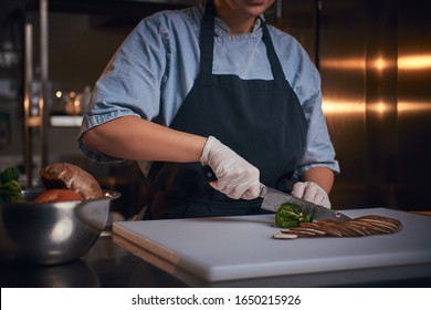 Chef female standing on a dark restaurant kitchen, wearing gloves, apron and denim shirt, cutting vegetables on a cutting board net to the bowl with raw vegetables, close up view