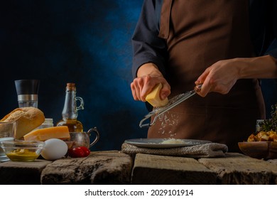 The chef grates the cheese for the Caesar salad. Cooking process. Lots of ingredients on a wooden table. Dark blue background. Healthy food, festive salad. Restaurant, home cooking. Close-up.