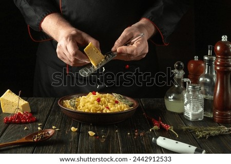 The chef grates hard cheese into a plate of pasta using a hand grater. Work environment in hotel kitchen with spices and kitchen utensils.