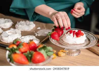 A chef in a green apron assembles strawberry pavlovas in a bright kitchen, with more desserts in soft focus behind. Ideal for culinary websites and dessert blogs. High quality photo - Powered by Shutterstock