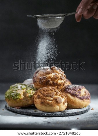 Chef hand dusting powdered sugar through a sieve over a stack of colorful mixed-flavor choux au craquelin. Action shot of dessert plating.
