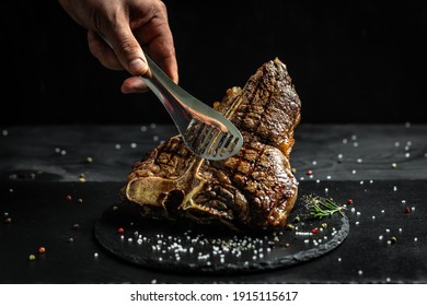 chef hand holding steak meat tongs. large piece of fresh beef meat prepared on a grill. Medium rare Grilled T-Bone Steak, Barbecue aged wagyu porterhouse.