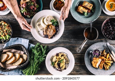 Chef Hand Picking Up A Plate With Meal From A Top View Of A Table Full Of Assortment Meals , Including Soup,bread,salad, Dumplings, Schnizel, Pasta Lasagne And Herbs.