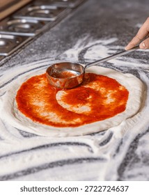 Chef hand is spreading pasteurized tomato paste onto a pizza base. The process of preparing pizza. Chef pours tomato sauce over the rolled dough.