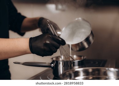 Chef hands in black gloves preparing food, whisking sauce or cream in a saucepan while pouring milk. Cooking process