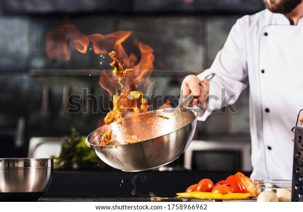 Chef hands\
keep wok with fire. Closeup chef hands cook food with fire. Chef\
man burn food at professional\
kitchen.