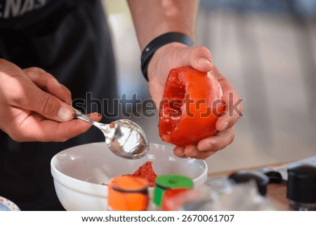 Chef hands scooping tomato pulp with metal spoon into white plate. Empty red tomatoes without flesh. Realistic candid kitchen scene of food preparation.