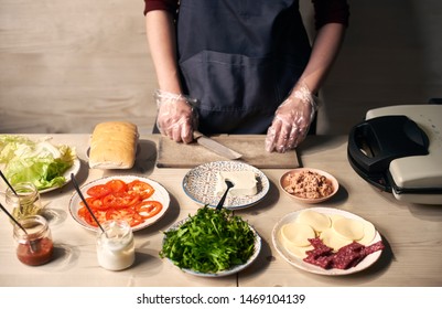 Chef Hands Taking Knife After Cutting Of Ingredients For Cooking Panini. Sandwich Grill Maker, Cheese, Salami, Tomato, Tuna, Butter, Bread Roll, Salad Leaves On Plates And Sauces On Light Wooden Table