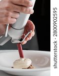 Chef hands using a culinary siphon to add gourmet foam over an appetizer on a white plate. Close-up, fine dining process.