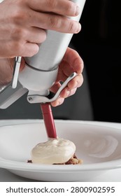 Chef hands using a culinary siphon to add gourmet foam over an appetizer on a white plate. Close-up, fine dining process.